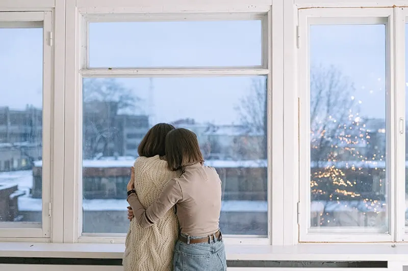 woman consoling her female friend hugging her near the window