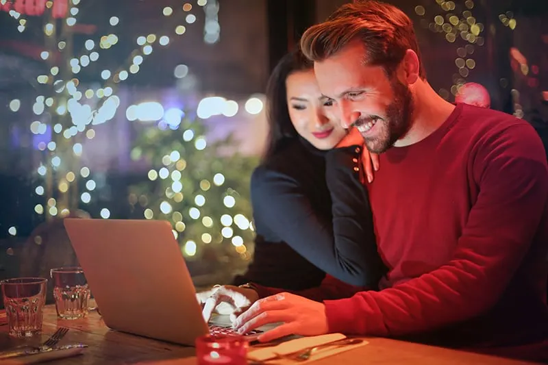 woman leaning on her smiling boyfriend while he is using laptop in a restaurant