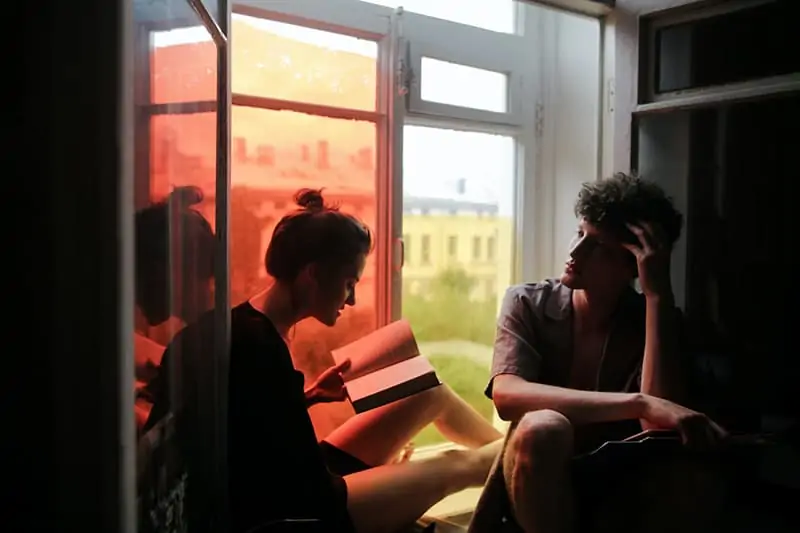 woman reading a book to her boyfriend while sitting on windowsill