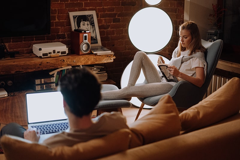 donna che legge un libro mentre il suo ragazzo usa il computer portatile in salotto la sera