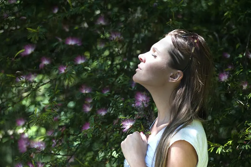 woman with closed eyes facing the sunlight standing near flower plant