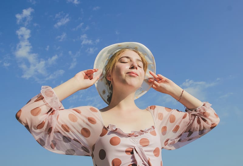 woman with closed eyes touching hat under blue sky