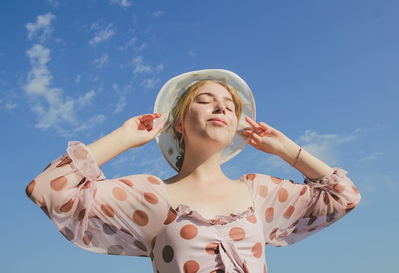 mujer con los ojos cerrados tocando el sombrero bajo el cielo azul