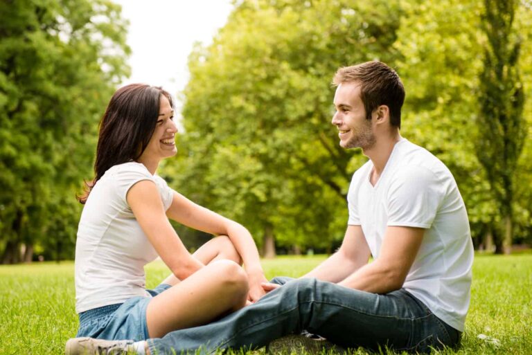 a smiling man and woman sitting on the grass holding hands and talking