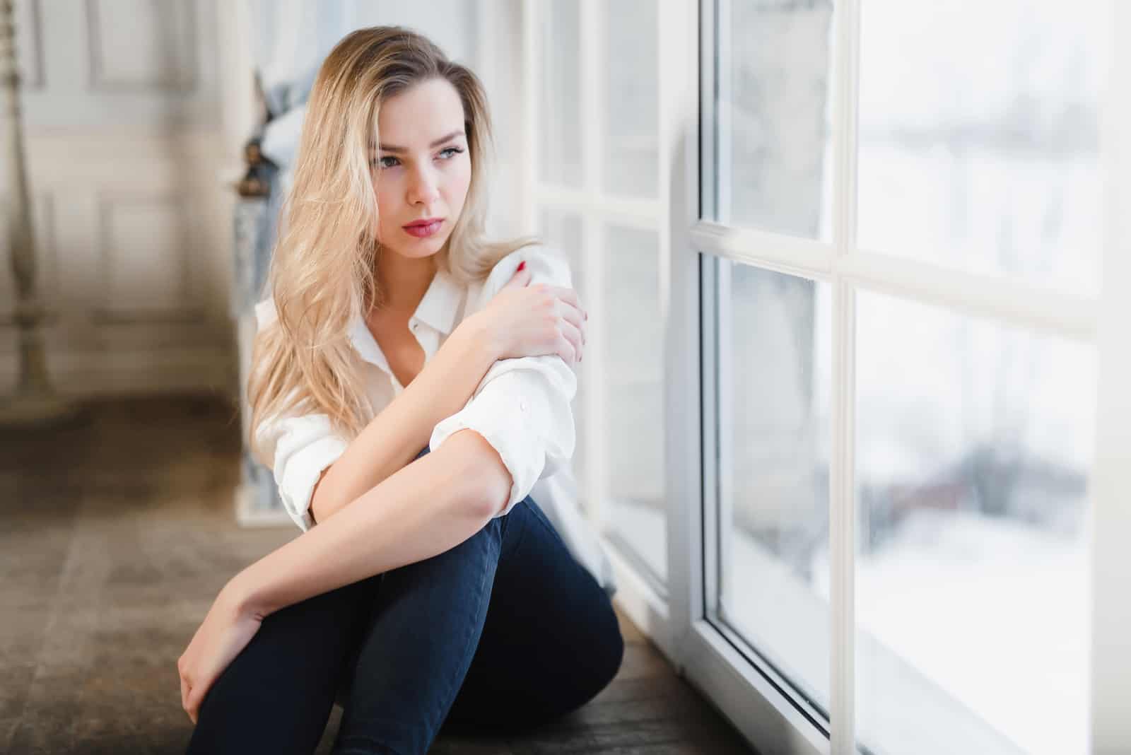 a sad woman with long blonde hair sits by the window