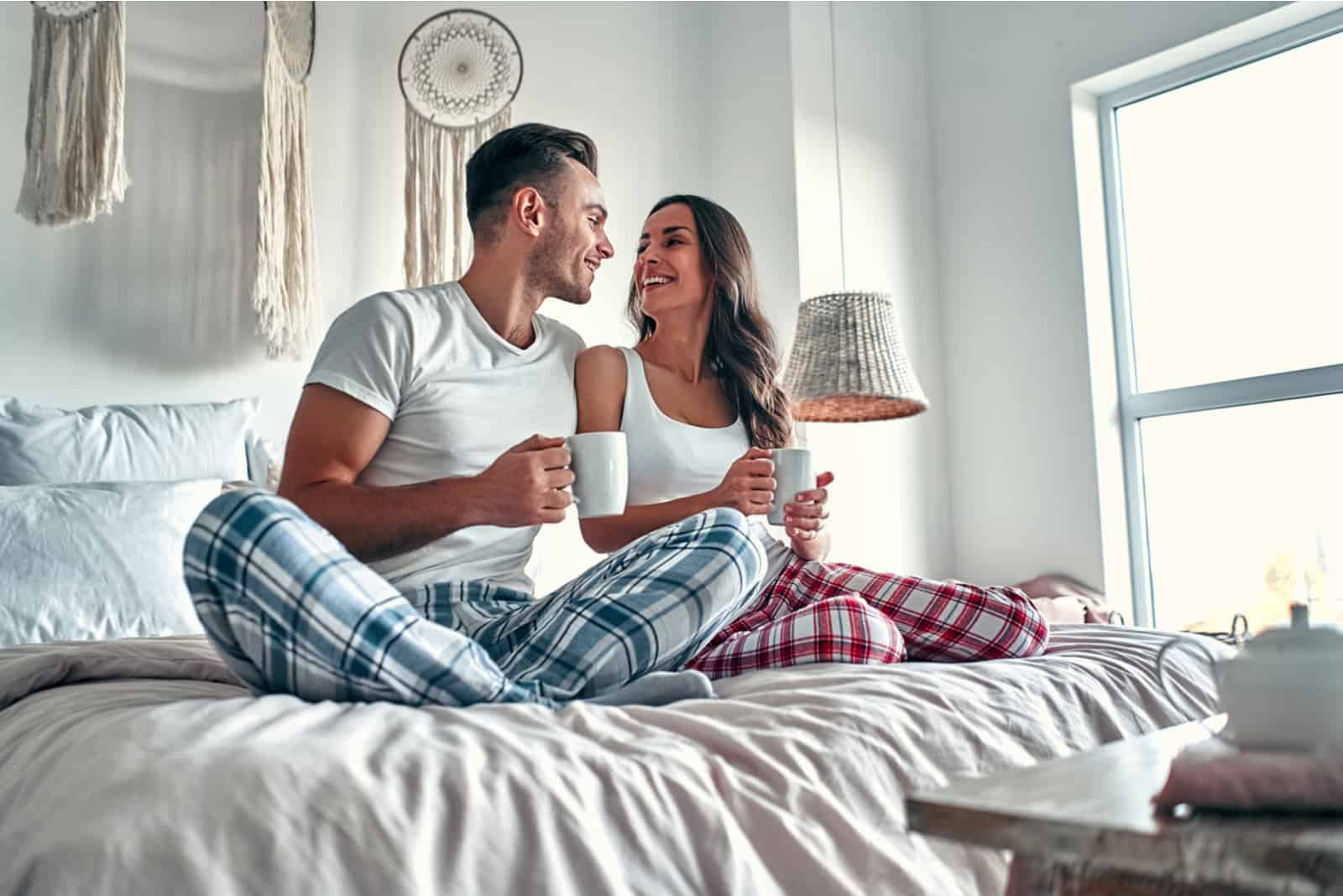 a smiling loving couple sitting on the bed and drinking coffee