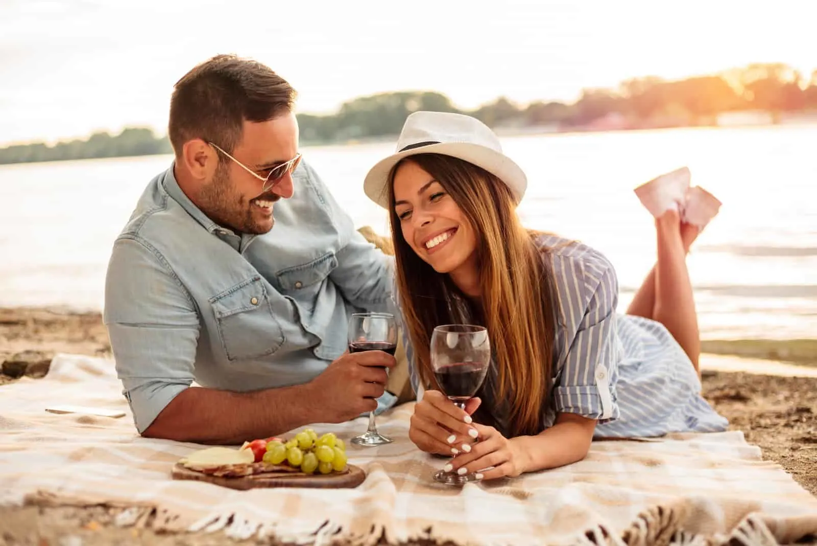a smiling man and woman lie on the beach and drink wine