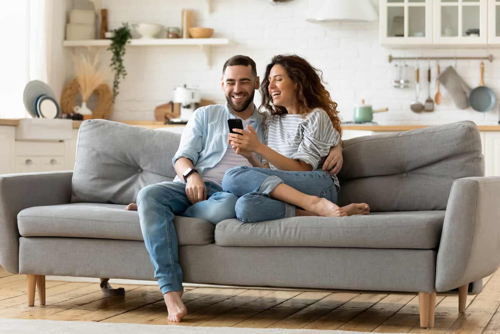 a smiling man and woman sit on the couch and look at the phone