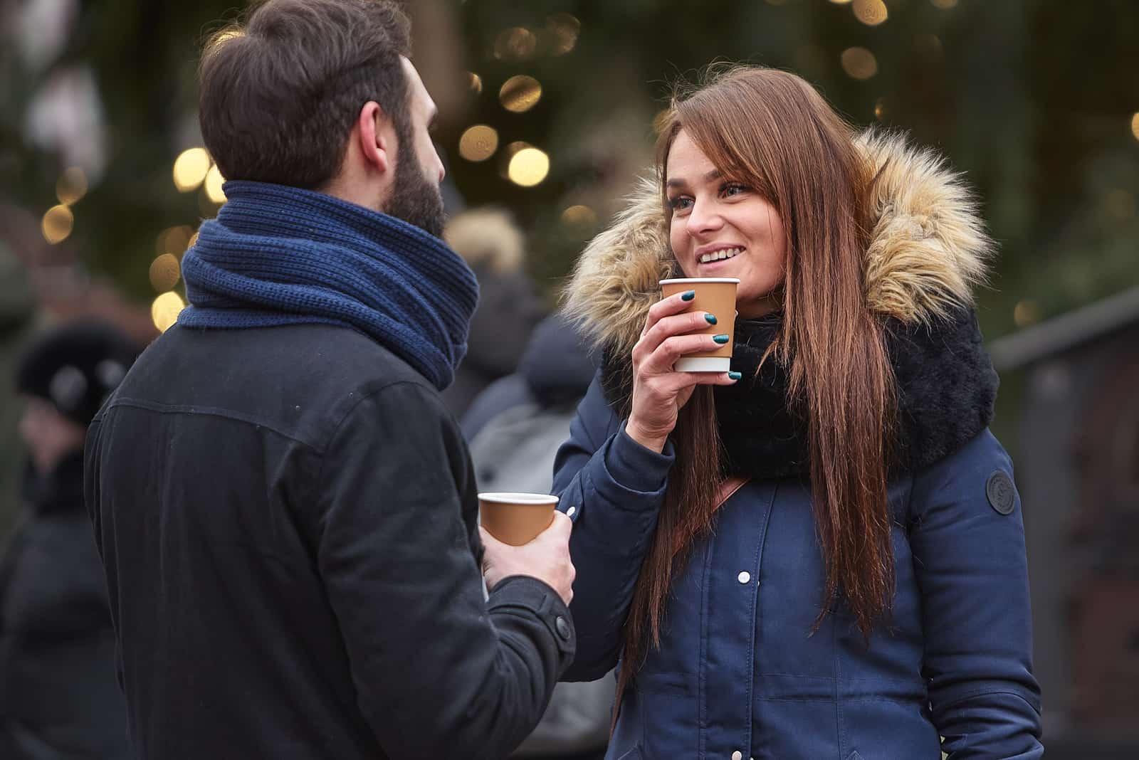 a smiling man and woman standing outdoors holding cups of coffee in their hand and talking