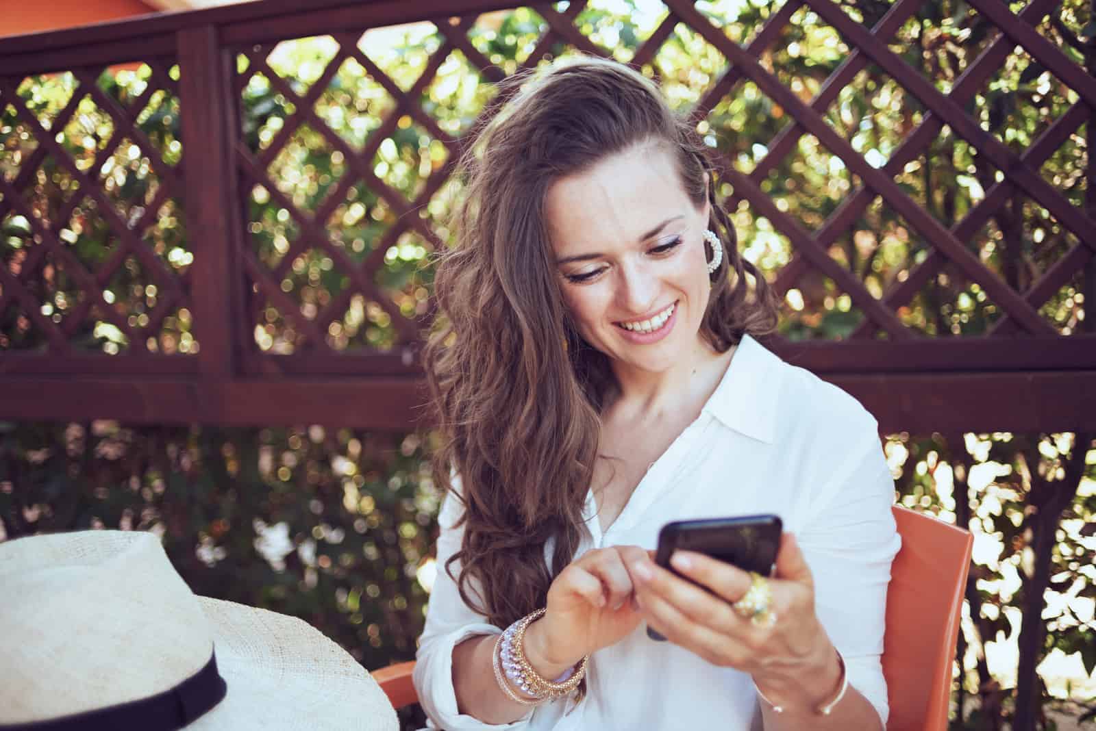 una mujer sonriente con el pelo largo y castaño se sienta en una mesa y teclea en el teléfono