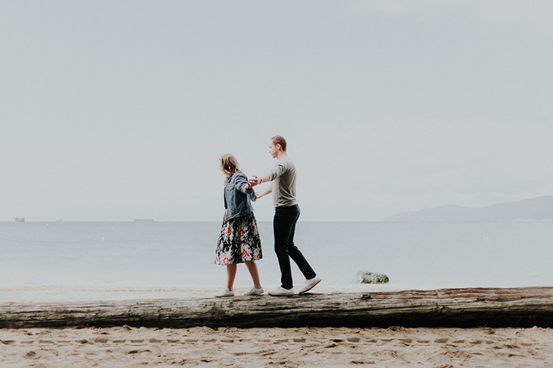 coppia che cammina sul tronco di legno sulla spiaggia e si tiene per mano