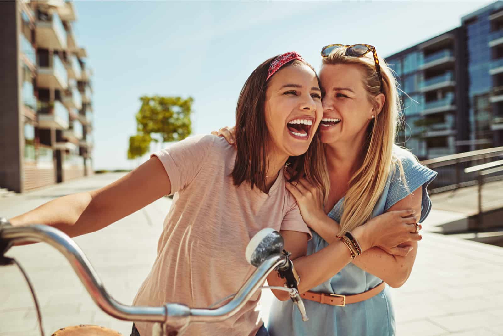 one girl rides a bike the other hugs and laughs