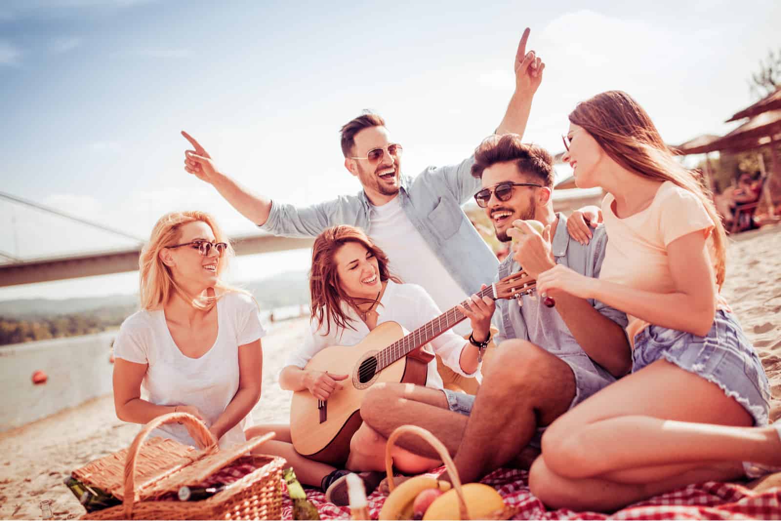 smiling friends sit on the beach and sing along to the guitar