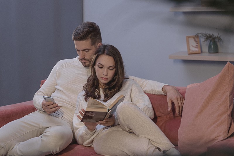 woman reading a book and man using his smartphone sitting on the couch close to each other