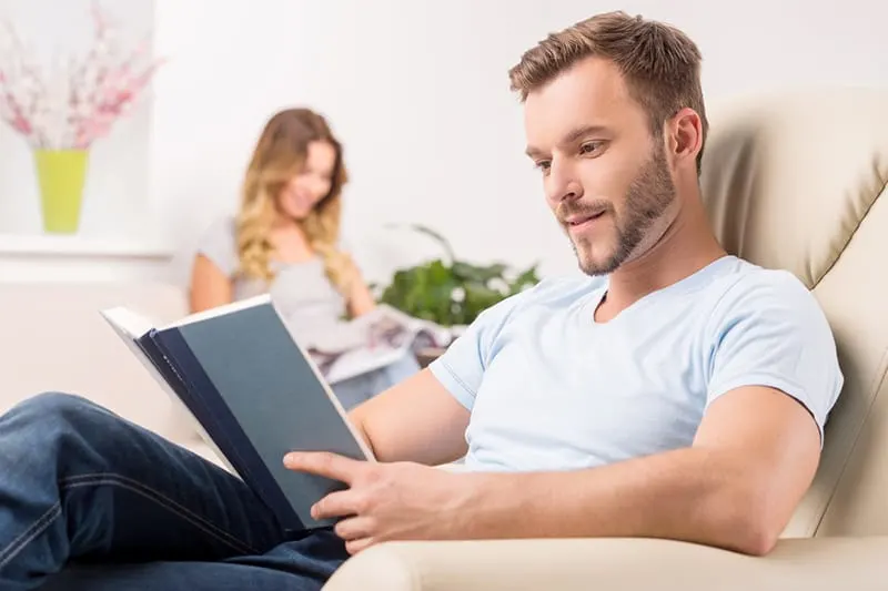  Cheerful young man reading book while his girlfriend sitting behind him and reading magazine