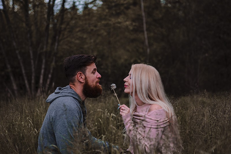 man and woman sitting in the field and blowing on dandelion