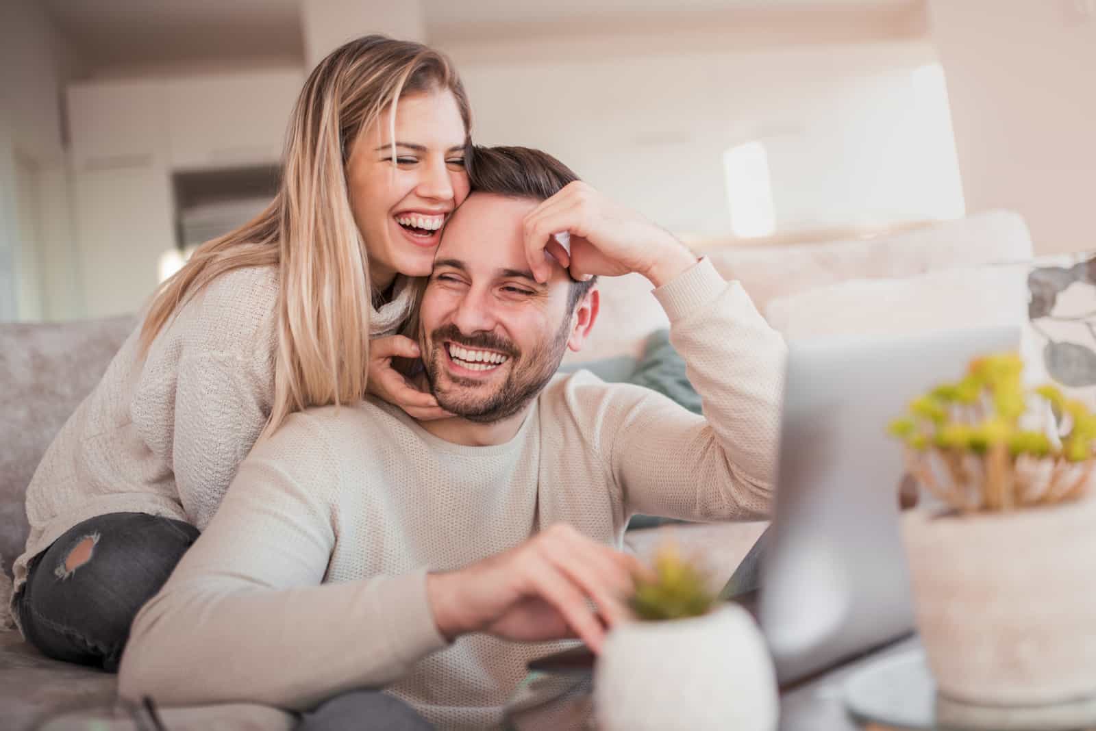 a man sitting behind a laptop a woman hugs him and laughs
