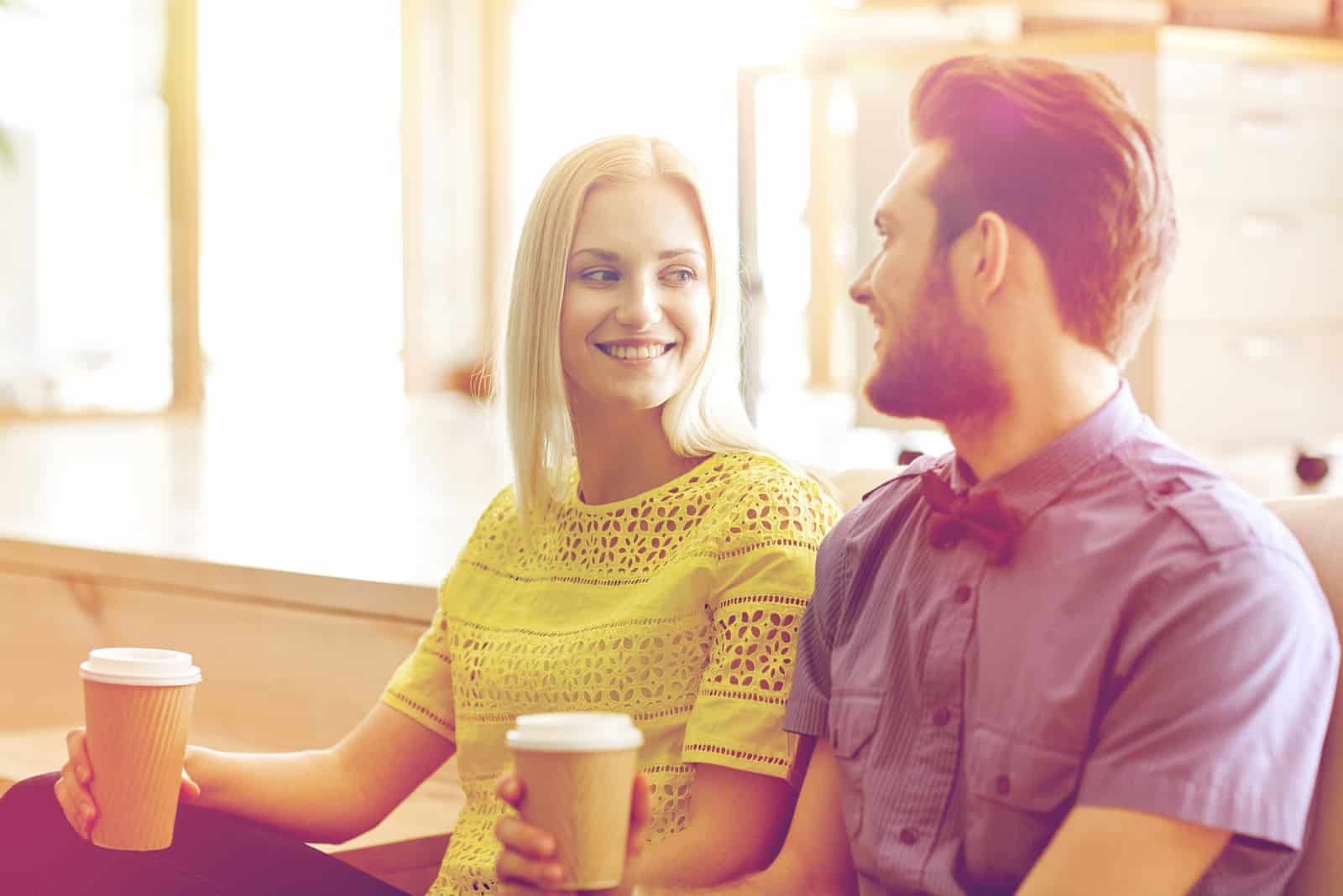 a smiling blonde-haired woman drinks coffee and talks to a man