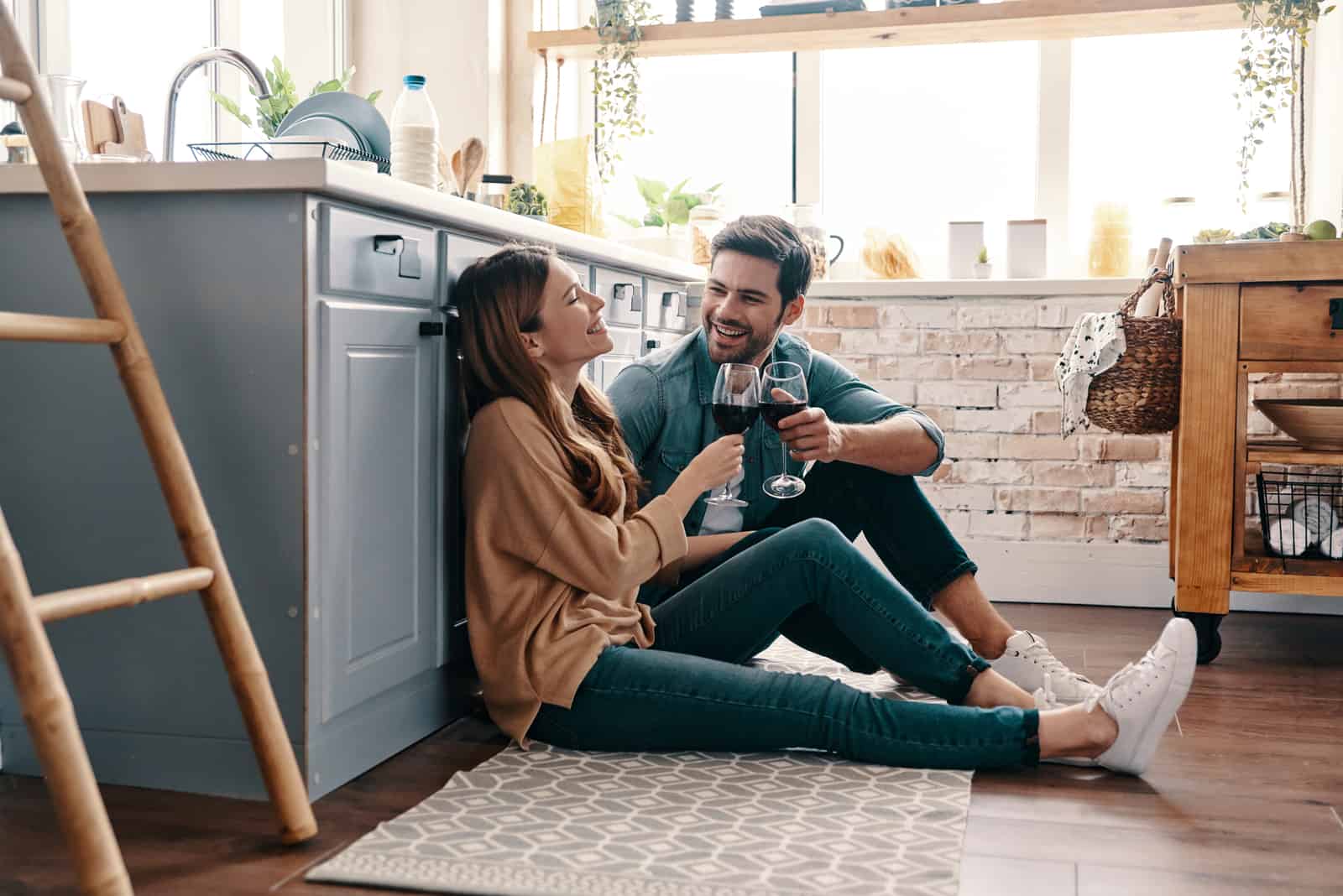 a smiling man and woman sit on the floor and drink wine