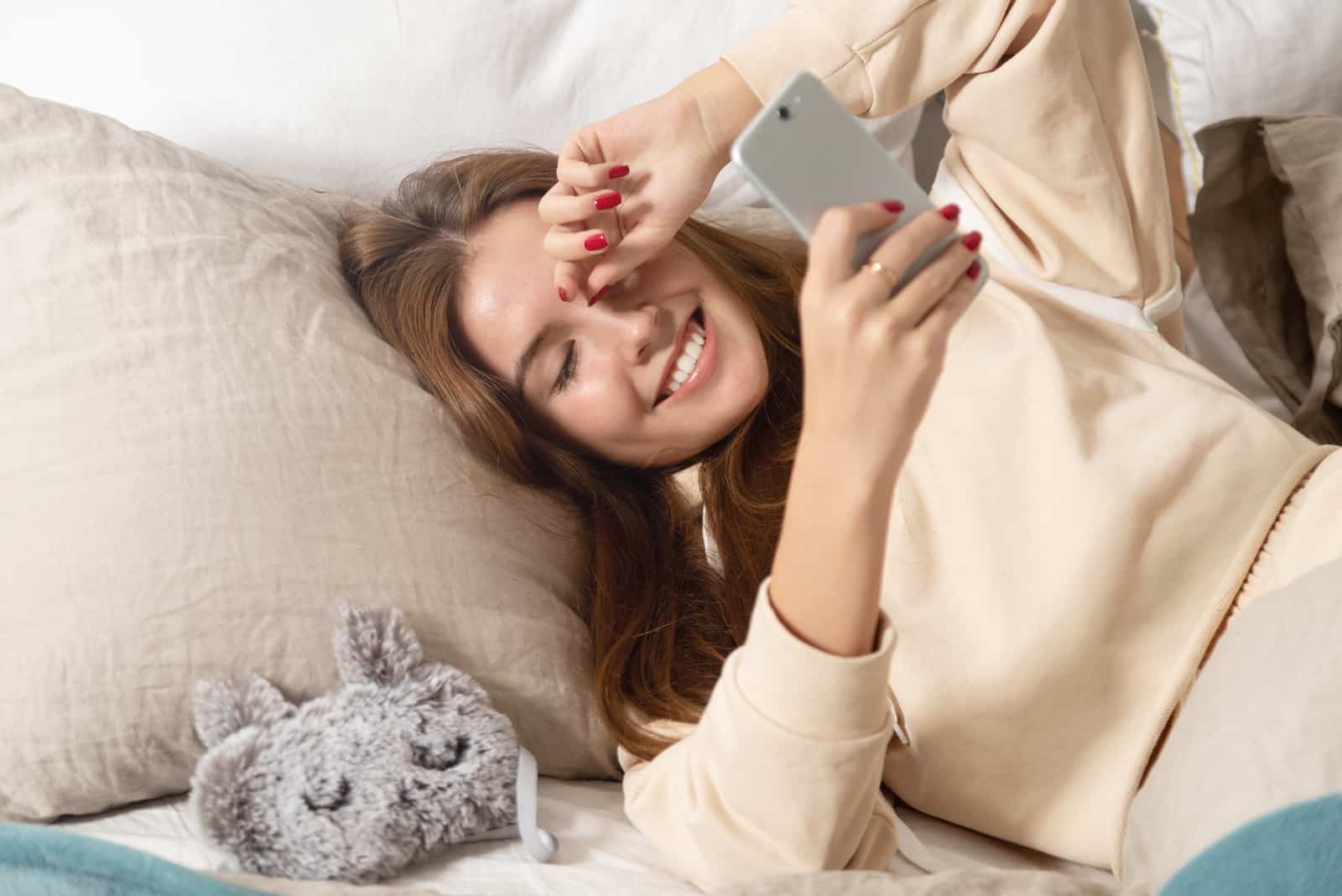 a smiling woman lies in bed and looks at the phone
