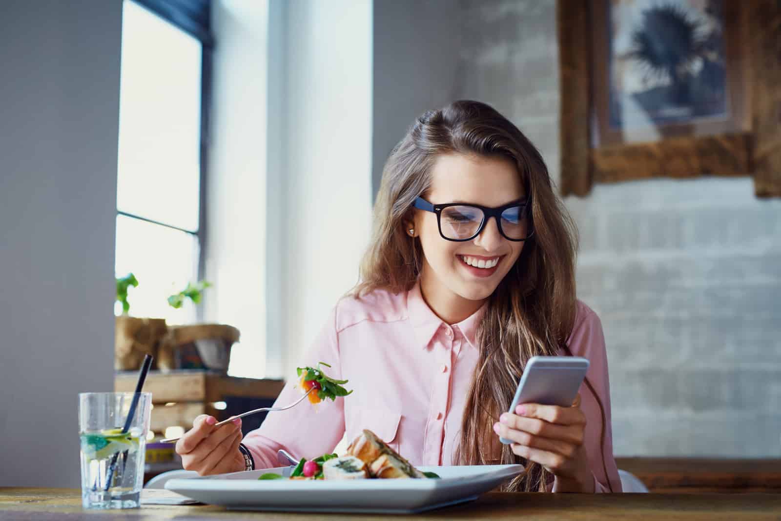 a smiling woman sitting at a table eating and pressing a phone