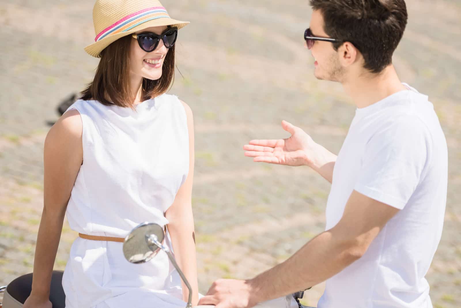 a smiling woman with a hat on her head sits on a motorcycle and talks to a man