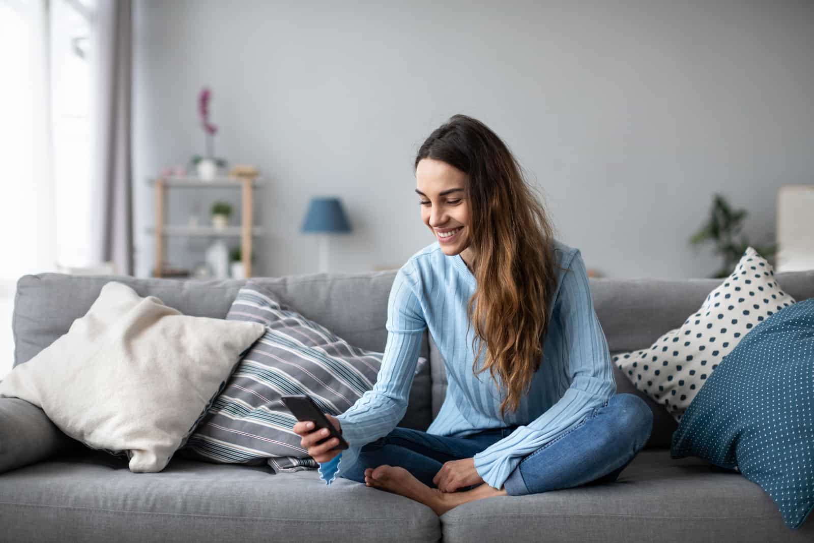 a smiling woman with long brown hair sits on the couch and keys on the phone