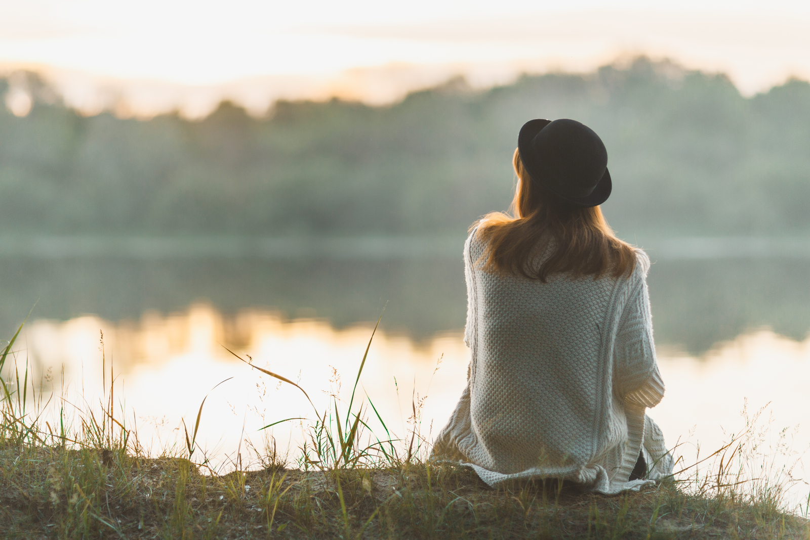 a woman with a hat on her head sits on the grass and looks out at the lake