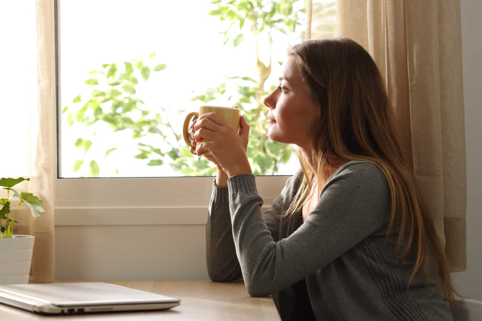 una donna con lunghi capelli castani siede a un tavolo bevendo caffè e guardando fuori dalla finestra