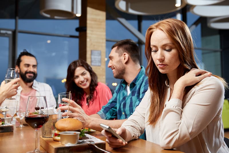 mujer aburrida cenando con amigos y enviando mensajes por smartphone en un restaurante