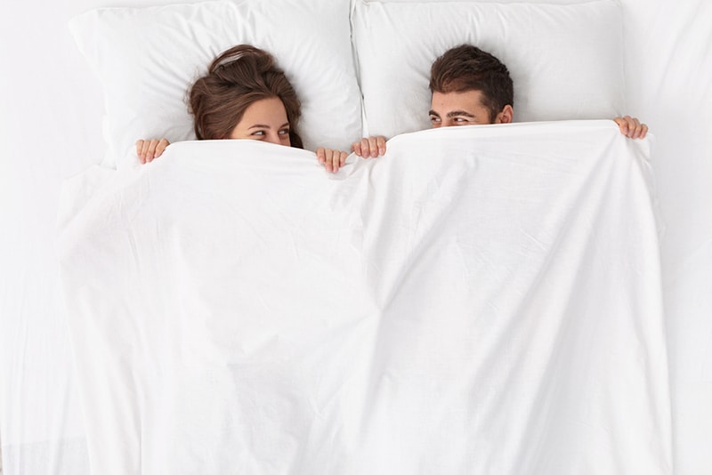 couple lying in the bed covered with white sheet and looking at each other