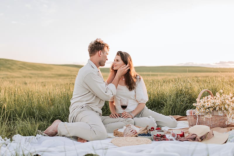 couple sitting on the blanket in the field and looking each other