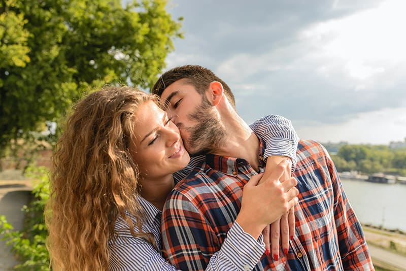 man kissing his girlfriend on the cheek while she embracing him from the back