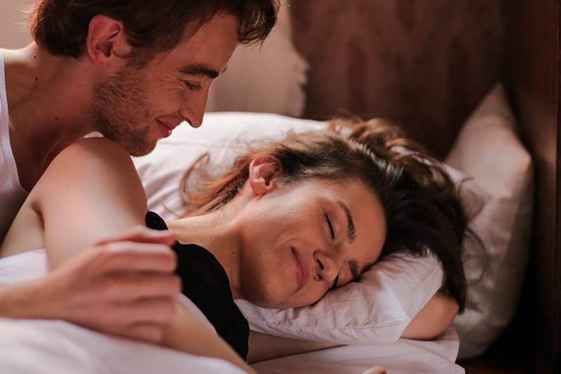 smiling man looking at his sleepy girlfriend lying in the bed