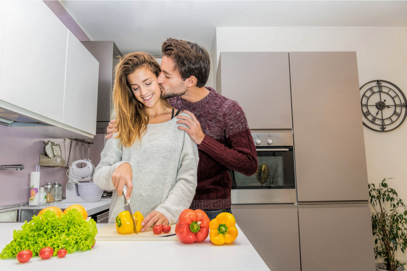 uma mulher sorridente corta legumes na cozinha e um homem beija-a na face