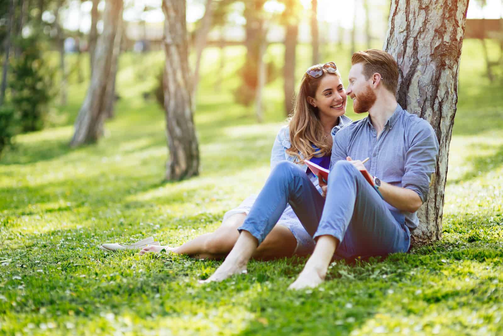 a man and a woman are sitting on the grass leaning against a tree and talking