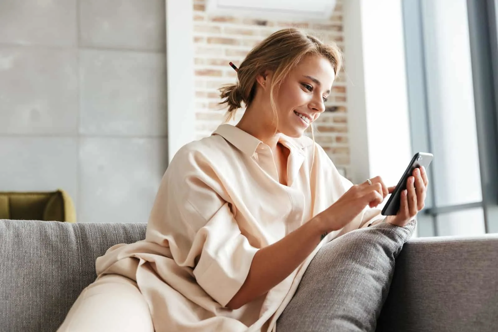a smiling blonde hair woman sits on the couch and keys on the phone