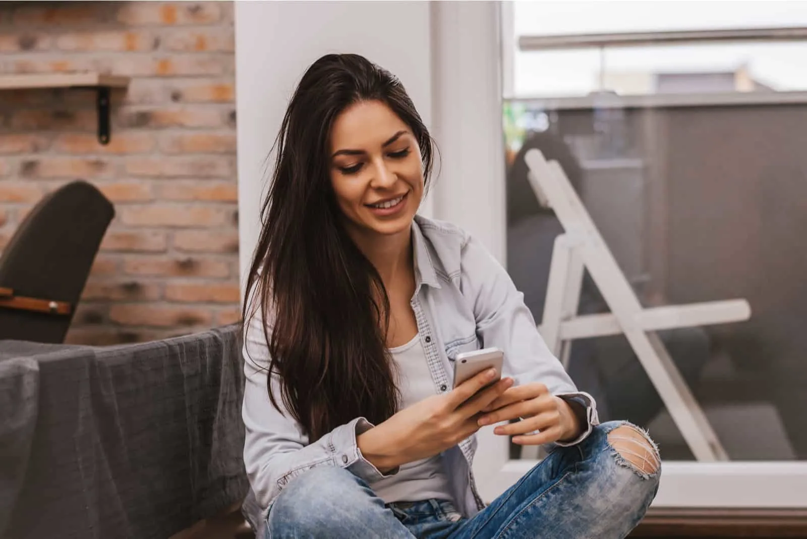 a smiling woman is sitting on the couch and typing on the phone