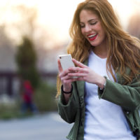 smiling brown haired woman standing on the street and button on the phone
