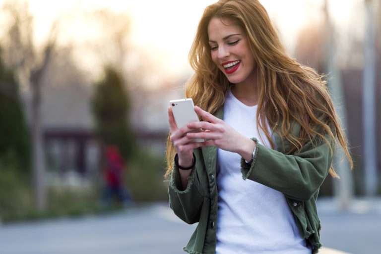 smiling brown haired woman standing on the street and button on the phone