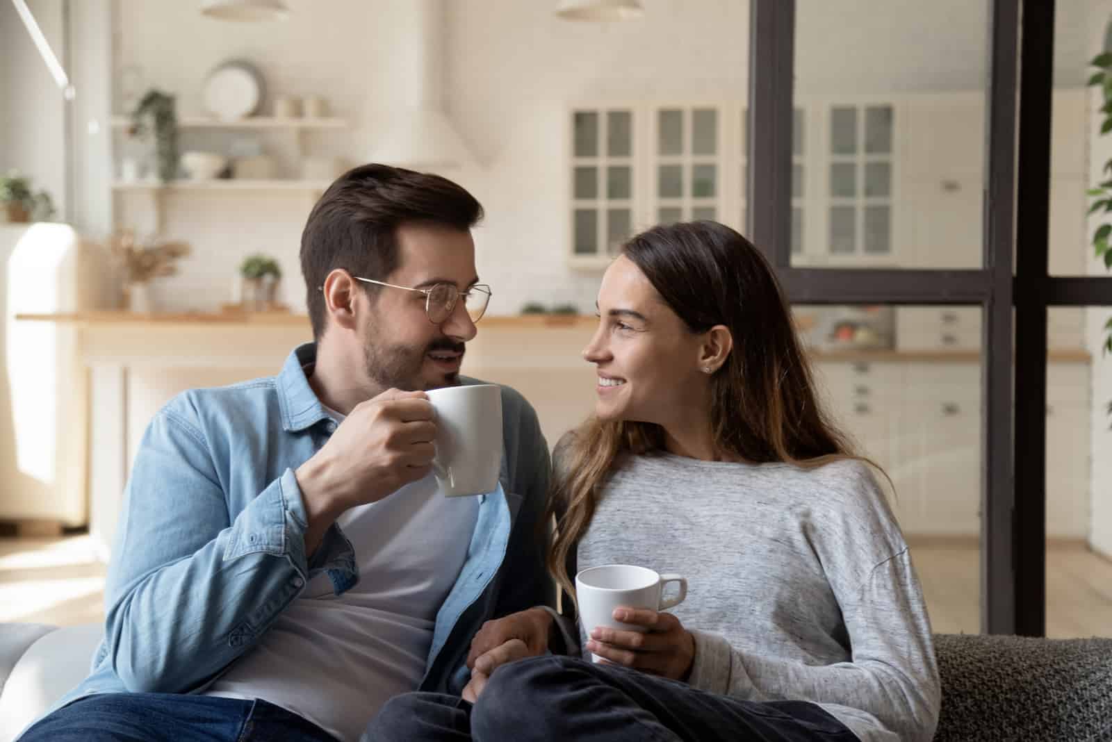 a smiling man and woman sit on the couch and talk over coffee