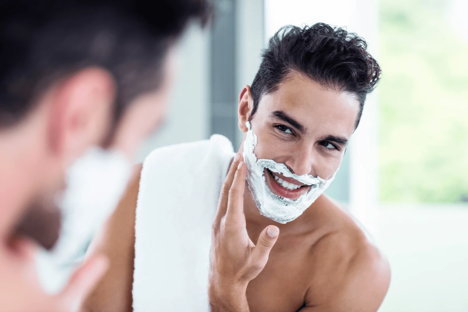 a smiling man stands in front of a mirror and shaves