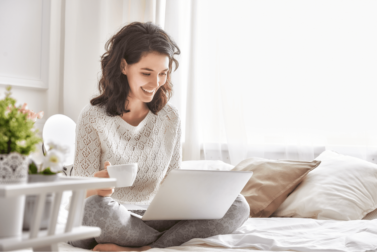 una donna sorridente siede dietro un computer portatile sul letto e tiene una tazza in mano