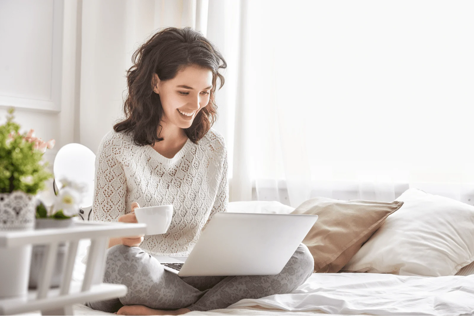 a smiling woman sits behind a laptop on the bed and holds a cup in her hand
