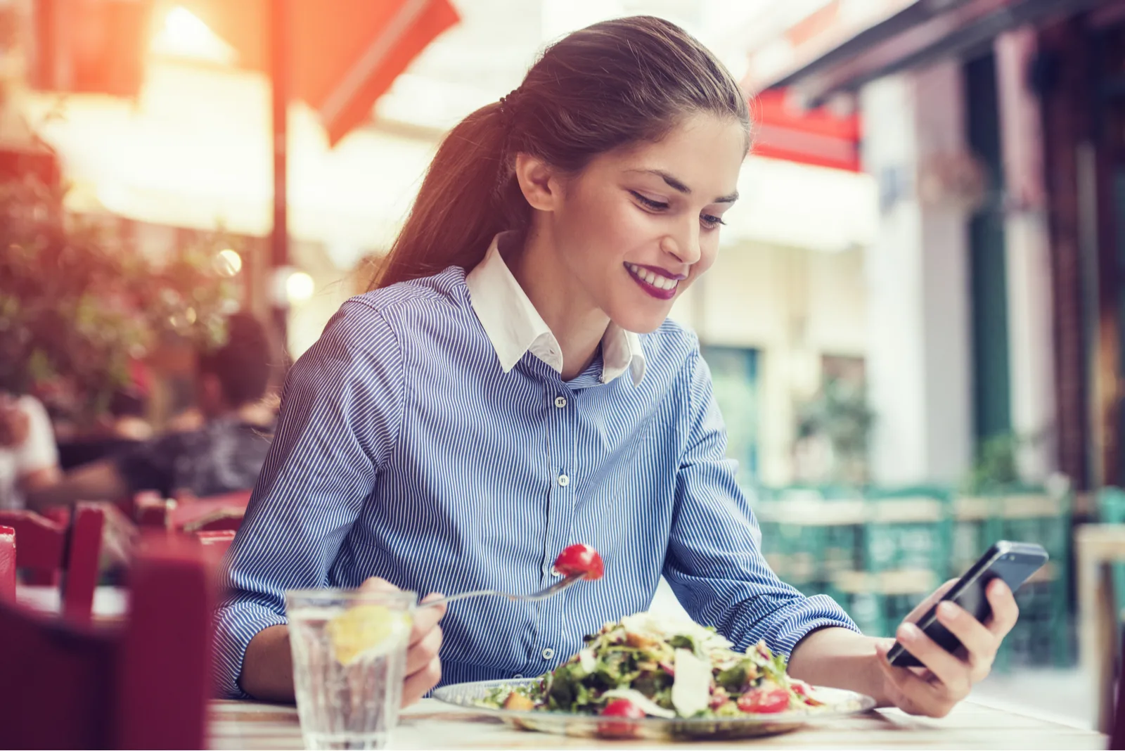 a smiling woman sitting at a table eating and holding a phone in her hand