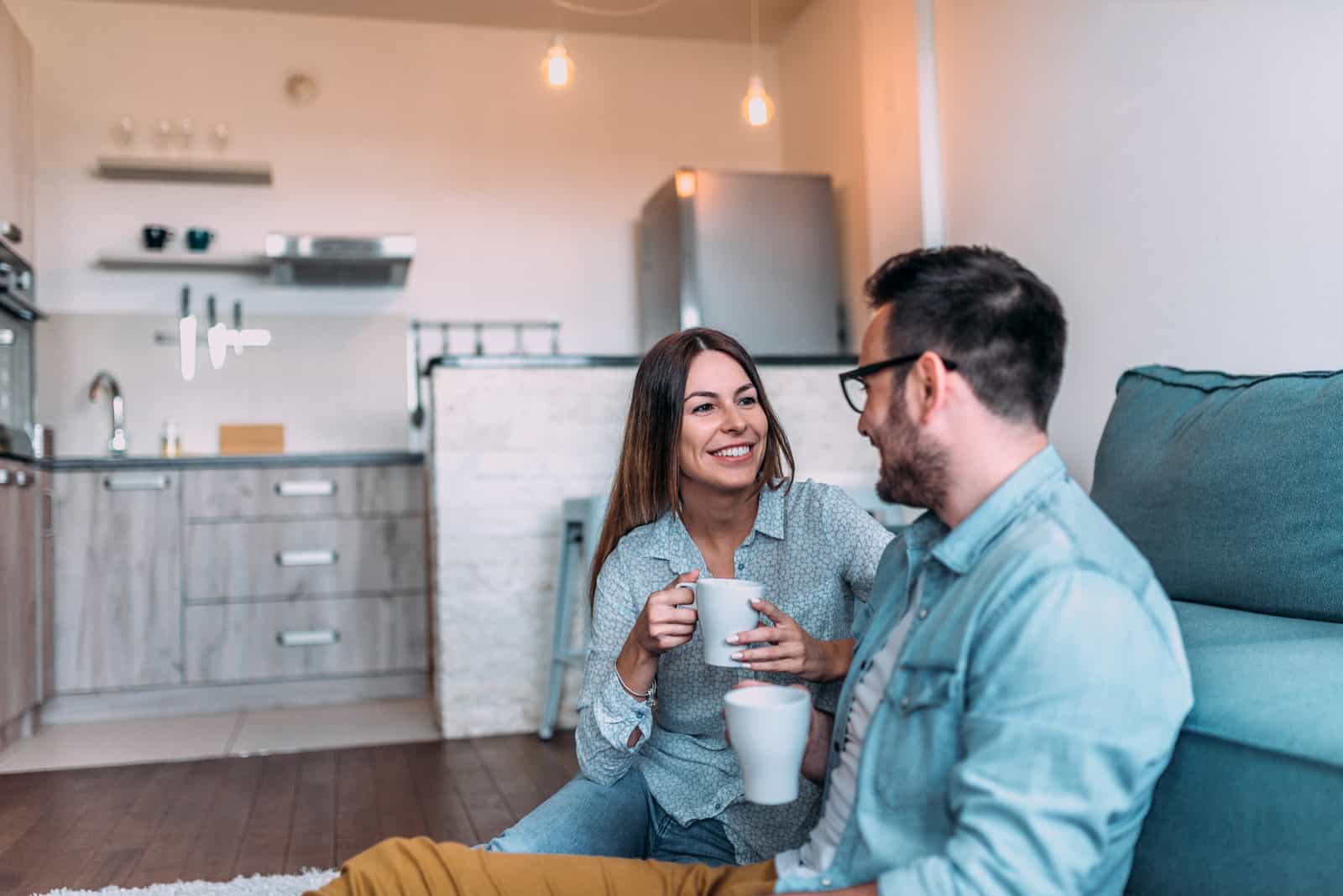 una coppia sorridente e innamorata seduta sul pavimento a bere caffè e a parlare