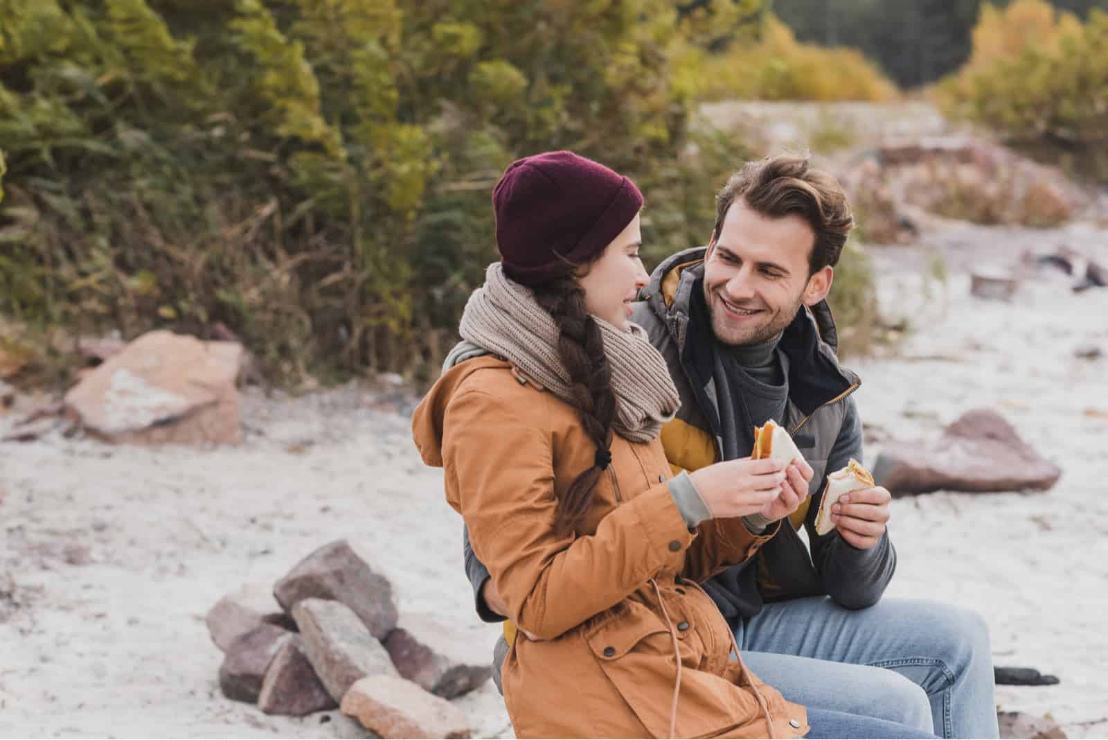 a smiling man sits next to a woman while holding a sandwich in his hands