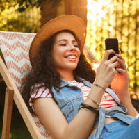 uma mulher sorridente com um chapéu na cabeça deitada numa cadeira e um botão no telefone