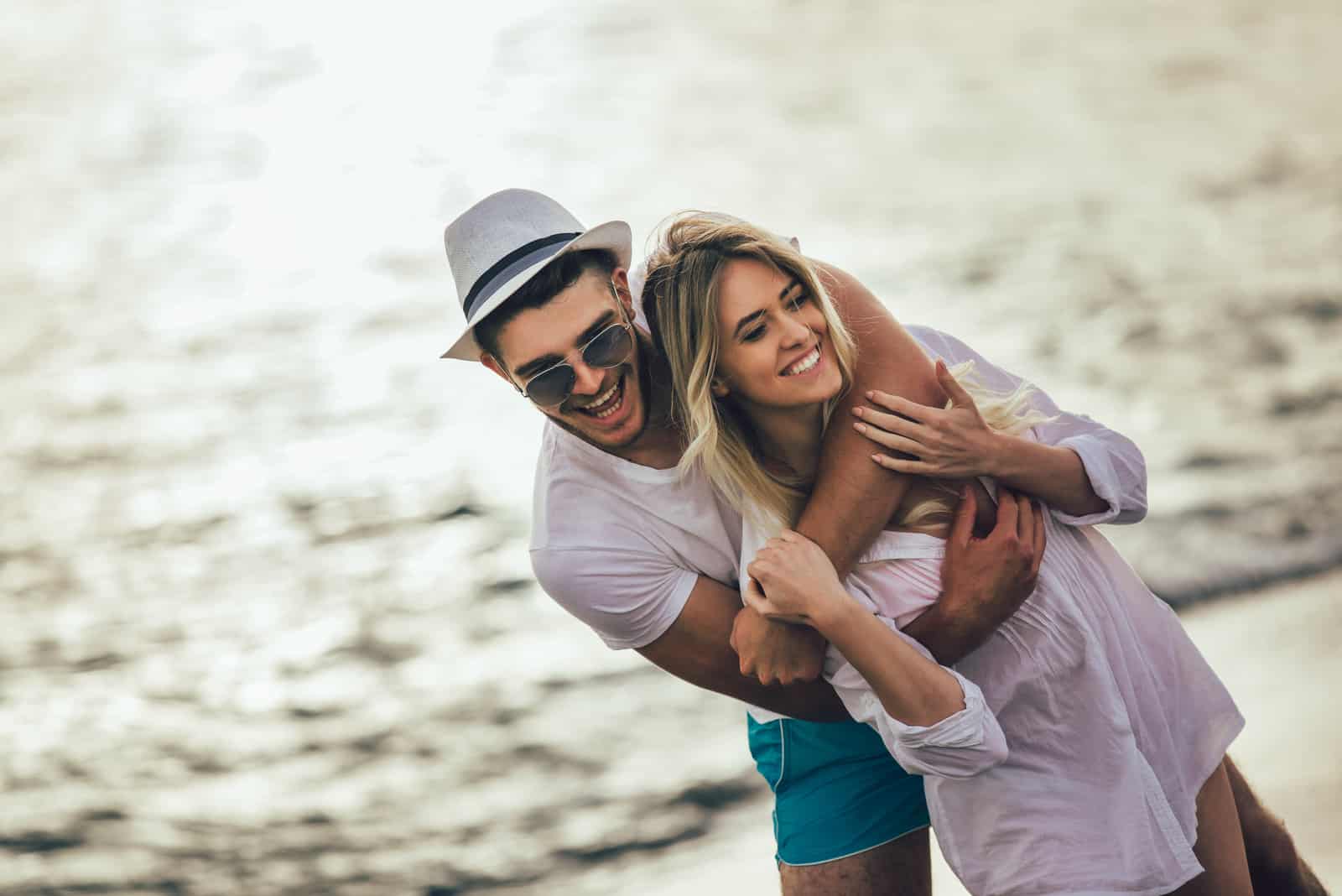 a smiling man with a hat on his head hugs a woman on the beach and laughs