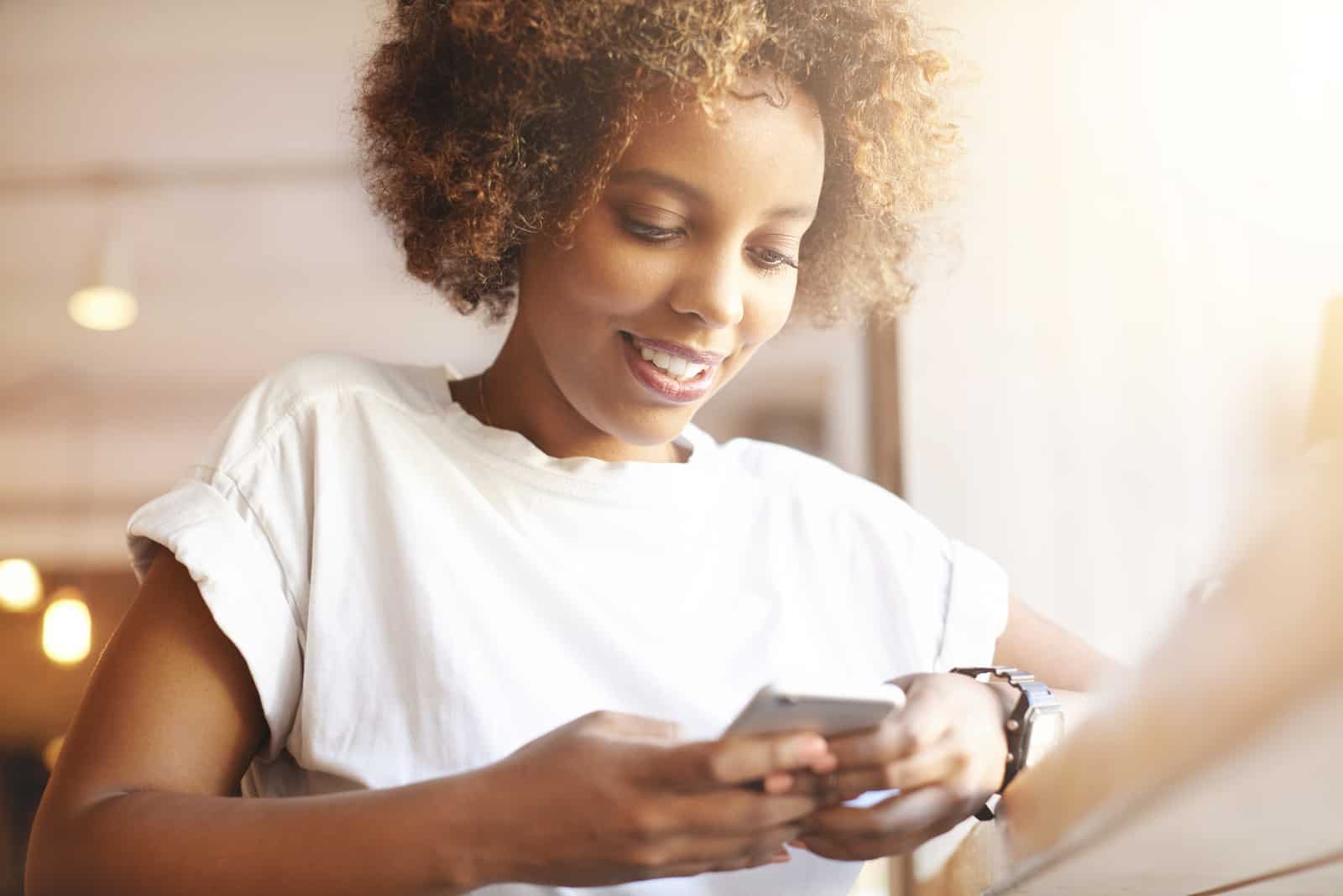 uma mulher sorridente com cabelo frisado sentada a uma mesa e a carregar no telemóvel